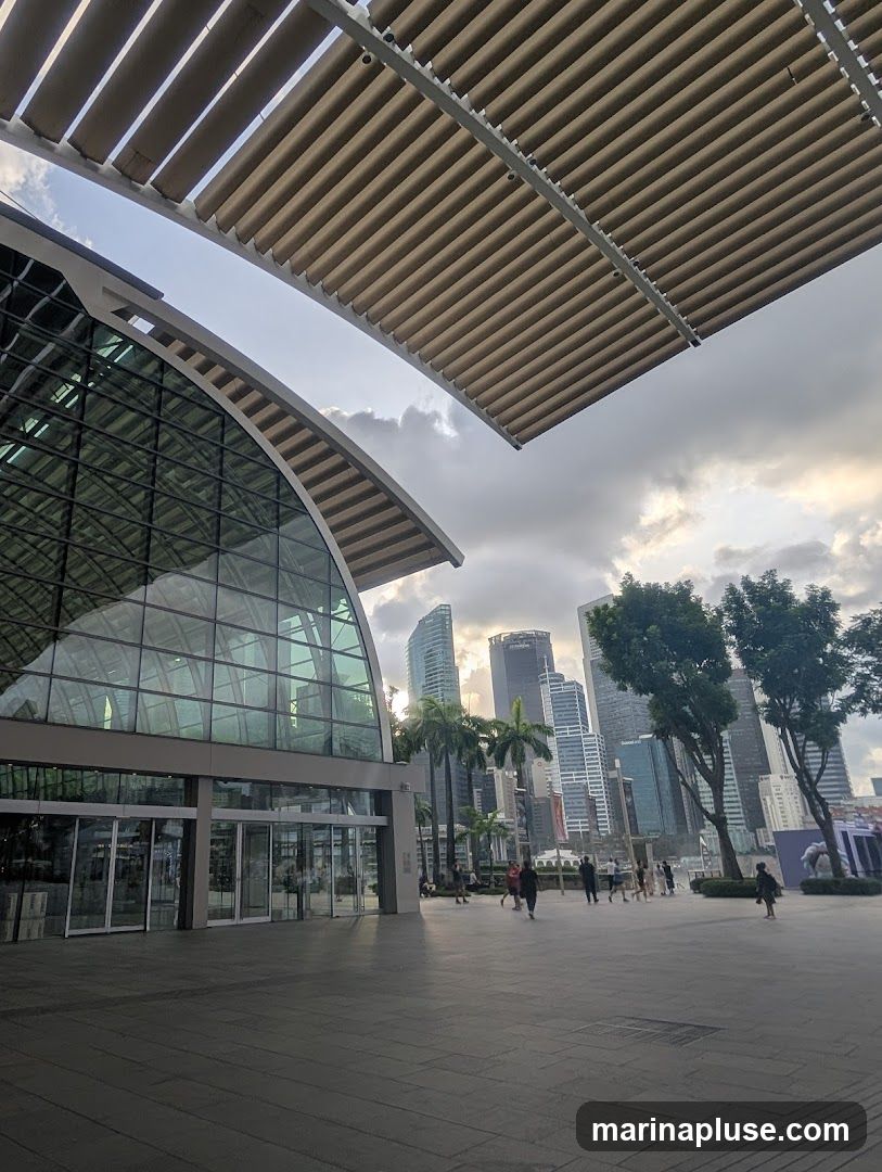 A wide shot of a modern building with a curved glass facade and a slatted roof structure. In the background, a city skyline with tall skyscrapers is visible under a cloudy sky. Palm trees and a few people are scattered on the paved plaza in front of the building.