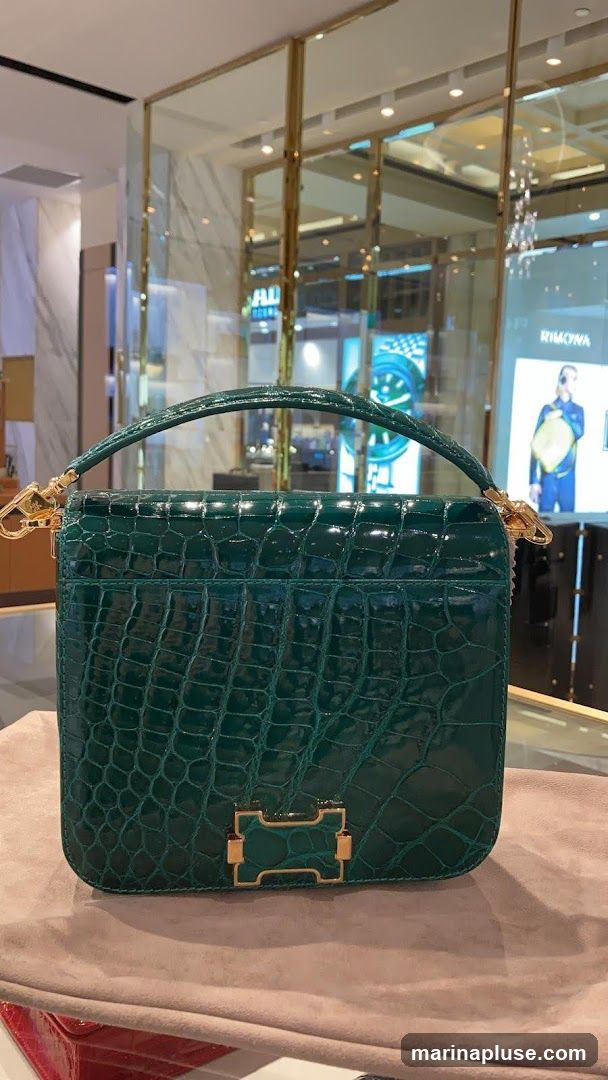 A close-up shot of a glossy green crocodile-skin handbag with gold hardware. The bag is displayed on a light brown velvet surface. In the background, a store interior is visible with glass displays and signage, including a prominent "RIMOWA" logo.