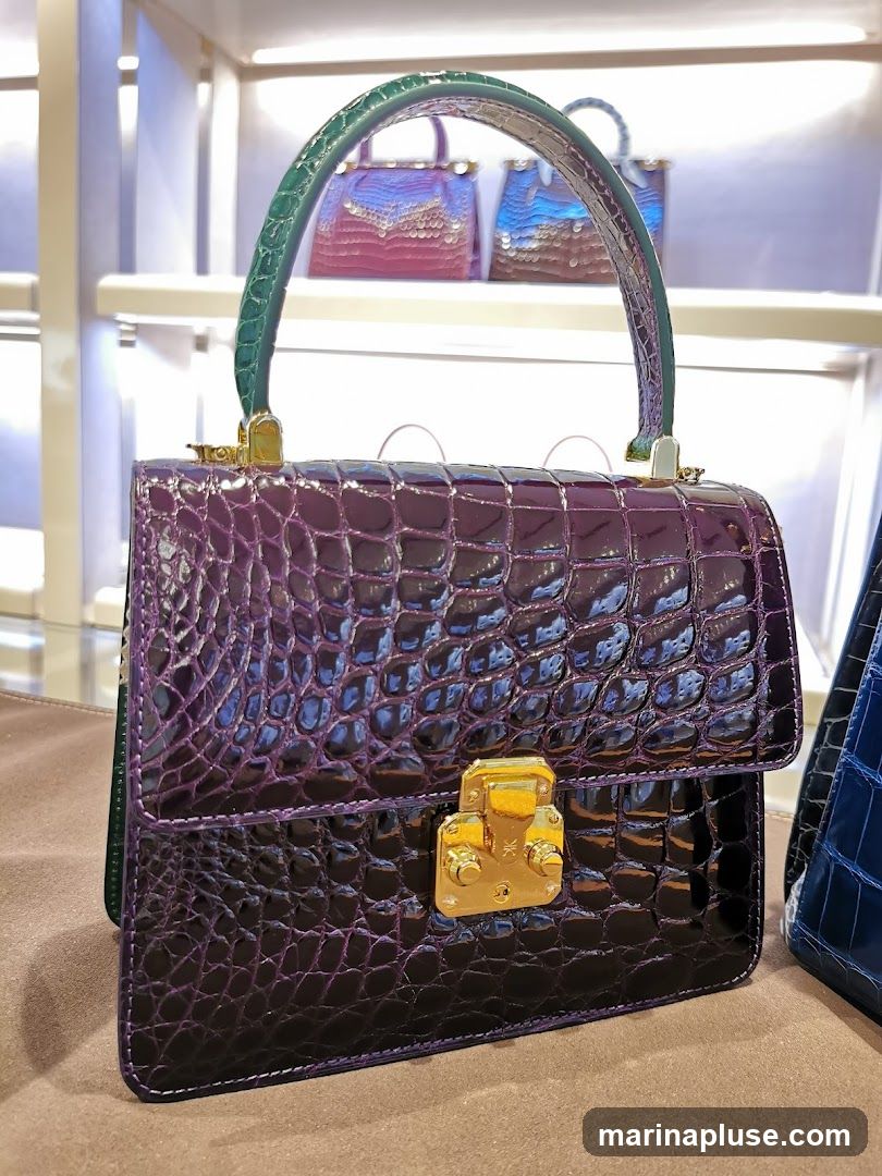 A close-up shot of a glossy purple crocodile handbag with a green handle and gold hardware. The bag is displayed on a brown surface, with other handbags visible in the blurred background on shelves.