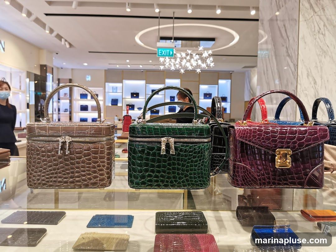 A display of various luxury handbags made of crocodile leather in different colors and styles. The handbags are arranged on shelves in a well-lit store. In the background, a store employee is visible, and shelves stocked with more bags are seen. An "EXIT" sign is also visible in the upper part of the image.