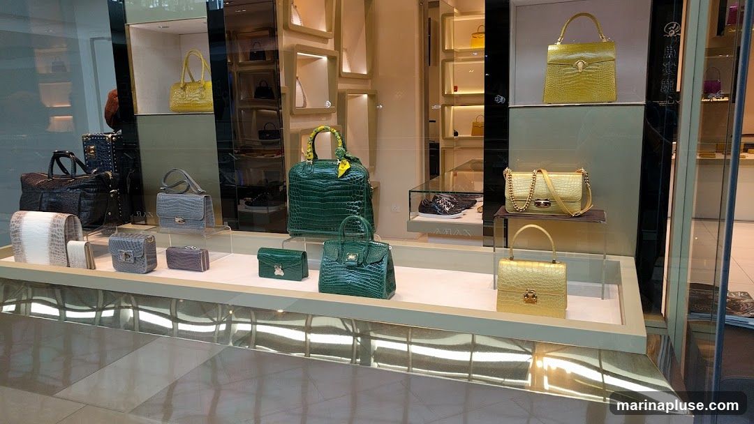 A well-lit display window showcases a collection of luxury handbags, primarily in crocodile-embossed leather. The bags are presented in various sizes and colors, including green, yellow, gold, grey, purple, and white. Some bags feature prominent gold hardware and chain straps. The display is organized on tiered shelves and within illuminated niches, creating an elegant and sophisticated presentation.