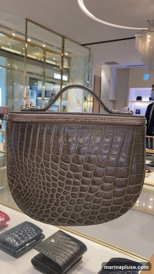 A close-up shot of a grey crocodile-embossed handbag with a top handle and zipper closure. Below the handbag, several wallets in black and blue, also with crocodile embossing, are displayed on a shelf. The background shows a brightly lit store interior with glass display cases and a person in a mask visible in the distance.