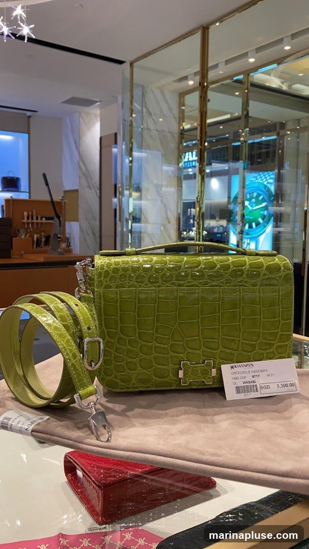 A close-up shot of a lime green crocodile handbag with a detachable strap. The bag is displayed on a beige cloth, with a smaller red clutch bag in the foreground. A price tag is attached to the handbag, indicating it is a crocodile handbag, model HBG CNF 8717 W21, color WASABI, priced at SGD 3,300.00. The background shows a well-lit store interior with display shelves and reflections.