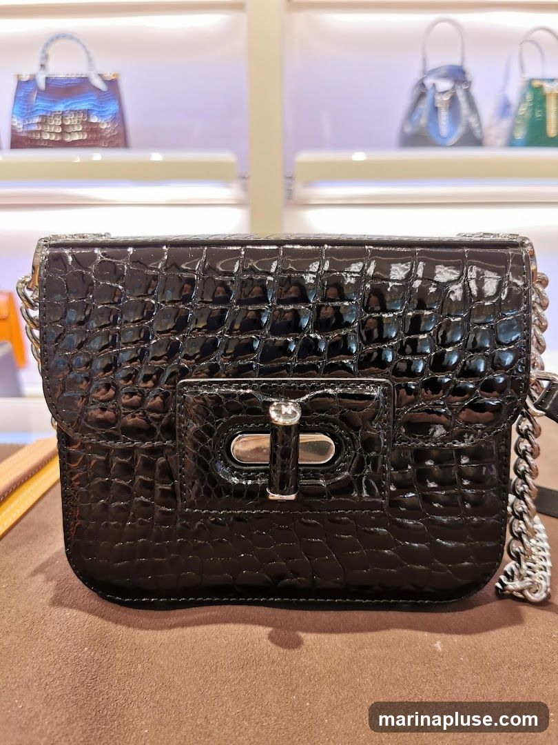 A close-up shot of a black crocodile-embossed leather handbag with a silver chain strap and a silver clasp. The handbag is displayed on a brown surface, with other handbags visible in the blurred background on shelves.