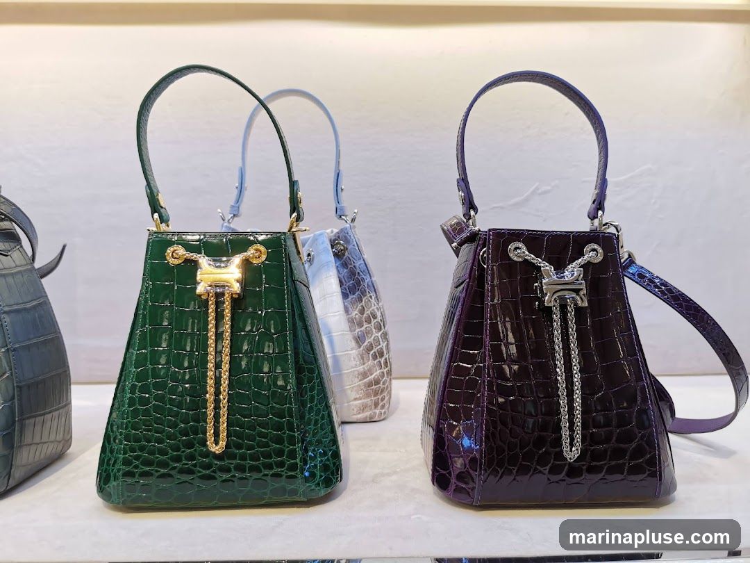 A display of several crocodile-embossed leather handbags in various colors, including green, purple, and a light grey/white. The bags have top handles and drawstring closures with decorative chain detailing. They are arranged on a shelf in what appears to be a retail setting.