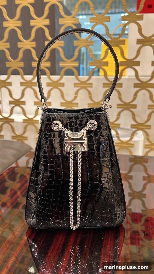 A black crocodile-textured handbag with silver hardware and chain detailing is displayed on a reflective surface. The background features a decorative gold pattern on a wall.