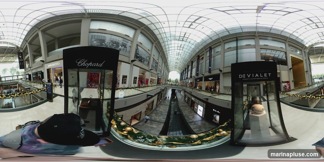 A panoramic view of the interior of a modern, multi-level shopping mall. The architecture features a large, arched glass ceiling allowing natural light to illuminate the space. Shops with prominent signage such as MIKIMOTO, Chopard, and DEVIALET are visible along the walkways. Decorative garlands with gold ornaments and greenery adorn the railings, suggesting a festive season. People are seen browsing and walking through the mall.