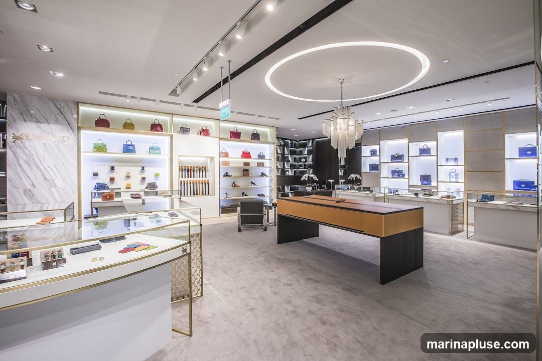 The interior of a high-end store, likely selling handbags and accessories. The store features a clean, modern design with marble accents, illuminated display shelves showcasing colorful handbags, and glass display cases with smaller items. A large, central display table with a leather top is prominent. The lighting is bright and sophisticated, with track lighting and a decorative chandelier. The overall ambiance is luxurious and elegant.