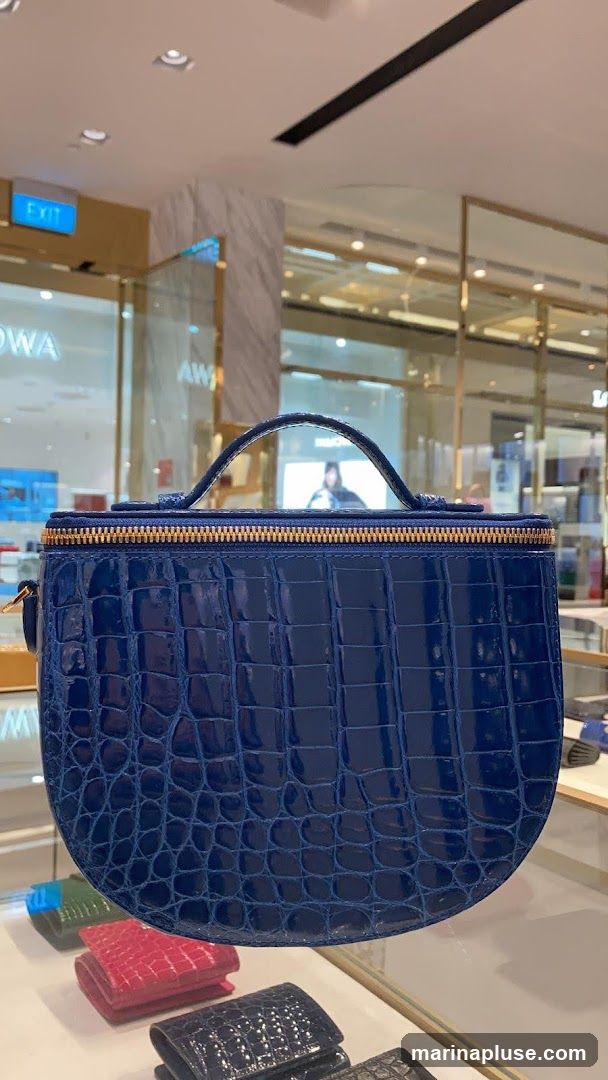 A close-up shot of a shiny blue crocodile-embossed handbag with a top handle and gold zipper. In the background, a retail store interior is visible with display shelves and an "EXIT" sign. Several smaller wallets in red, green, and black are displayed in the foreground.