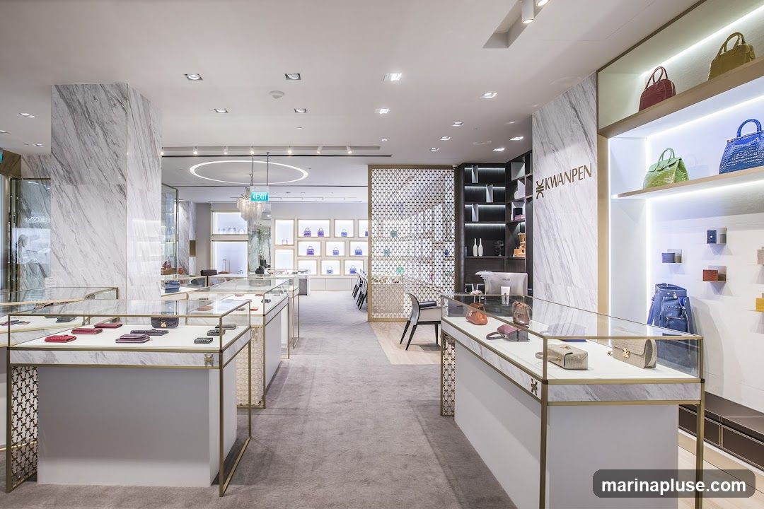 The interior of a high-end store displays various luxury handbags and accessories in illuminated display cases and on shelves. The store features marble accents, modern lighting, and a clean, minimalist aesthetic. A prominent logo for KWANPEN is visible on a marble pillar.