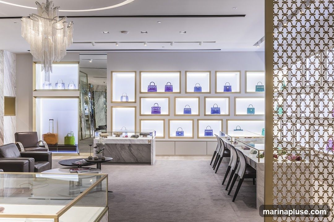 A well-lit, modern luxury store interior featuring a display wall with numerous handbags in various shades of blue and purple, each showcased in individual illuminated cubbies. To the left, a seating area with two leather armchairs and a coffee table is visible, along with a display of luggage and a green handbag. A long display counter runs along the right side of the room, with more handbags and accessories visible. A decorative screen with a geometric pattern is positioned on the far right. A large, ornate chandelier hangs from the ceiling.