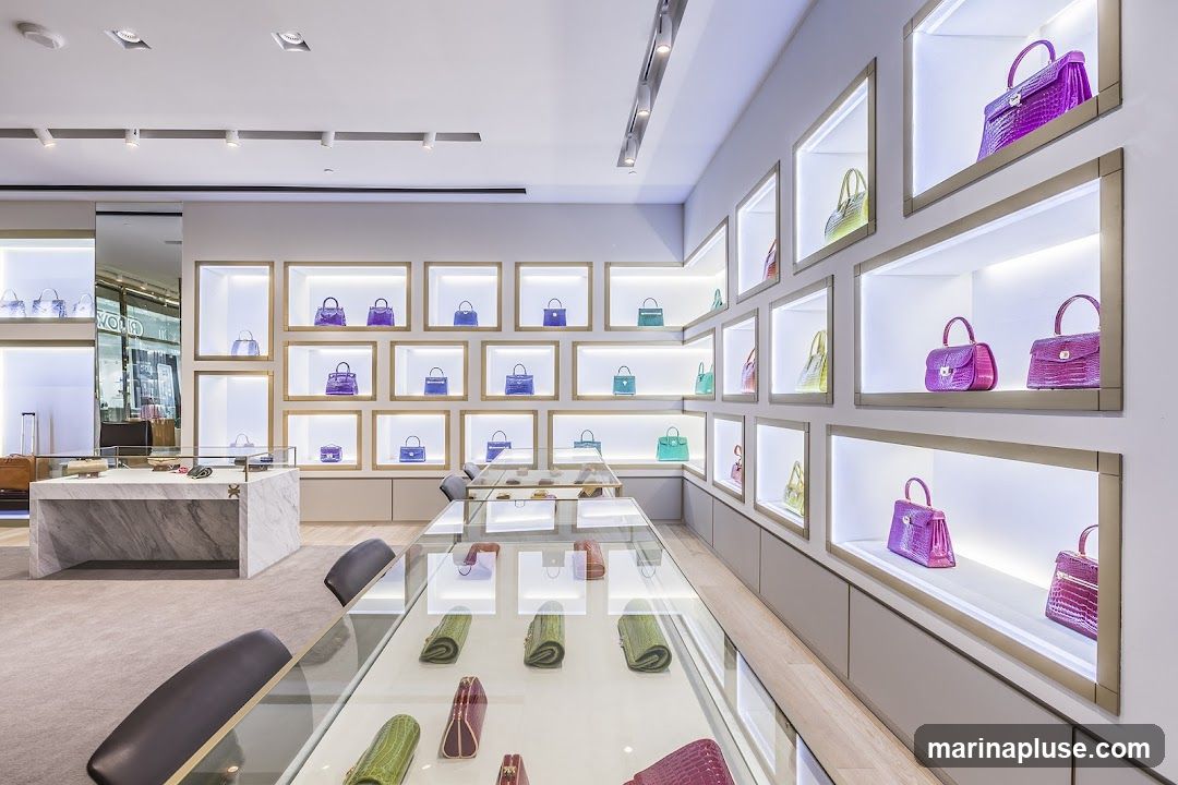 A brightly lit, modern interior of a luxury handbag store. Numerous handbags in various colors and styles are displayed in illuminated wall niches and glass display cases. The store features clean lines, marble accents, and plush seating.