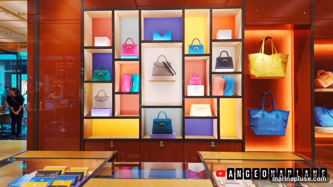 A retail display featuring a variety of handbags in different colors and styles, arranged on shelves with colorful backdrops. Some handbags are displayed in glass cases at the bottom. A YouTube logo and channel name are visible in the lower right corner.