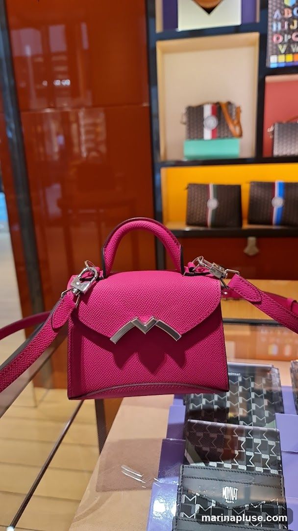 A close-up shot of a small, bright pink handbag with a silver clasp and handle. The bag is displayed on a shelf in what appears to be a retail store. In the background, other luxury accessories like wallets and small bags are visible on shelves, along with decorative elements and shelving units. The overall impression is of a high-end boutique display.