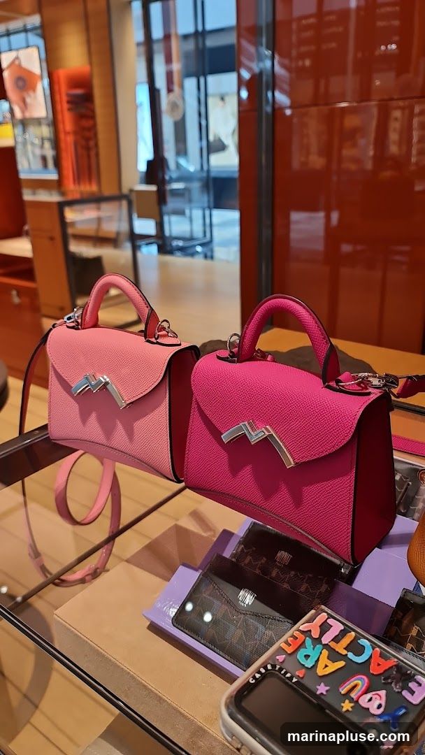 Two small, structured designer handbags are displayed on a glass shelf. The handbag on the left is a lighter pink, and the handbag on the right is a brighter fuchsia. Both bags feature a silver metallic clasp in the shape of a stylized 'M' or 'W'. Below the handbags, several cardholders and a phone case with colorful lettered stickers are visible. The background shows a retail store interior with displays and reflections in glass.