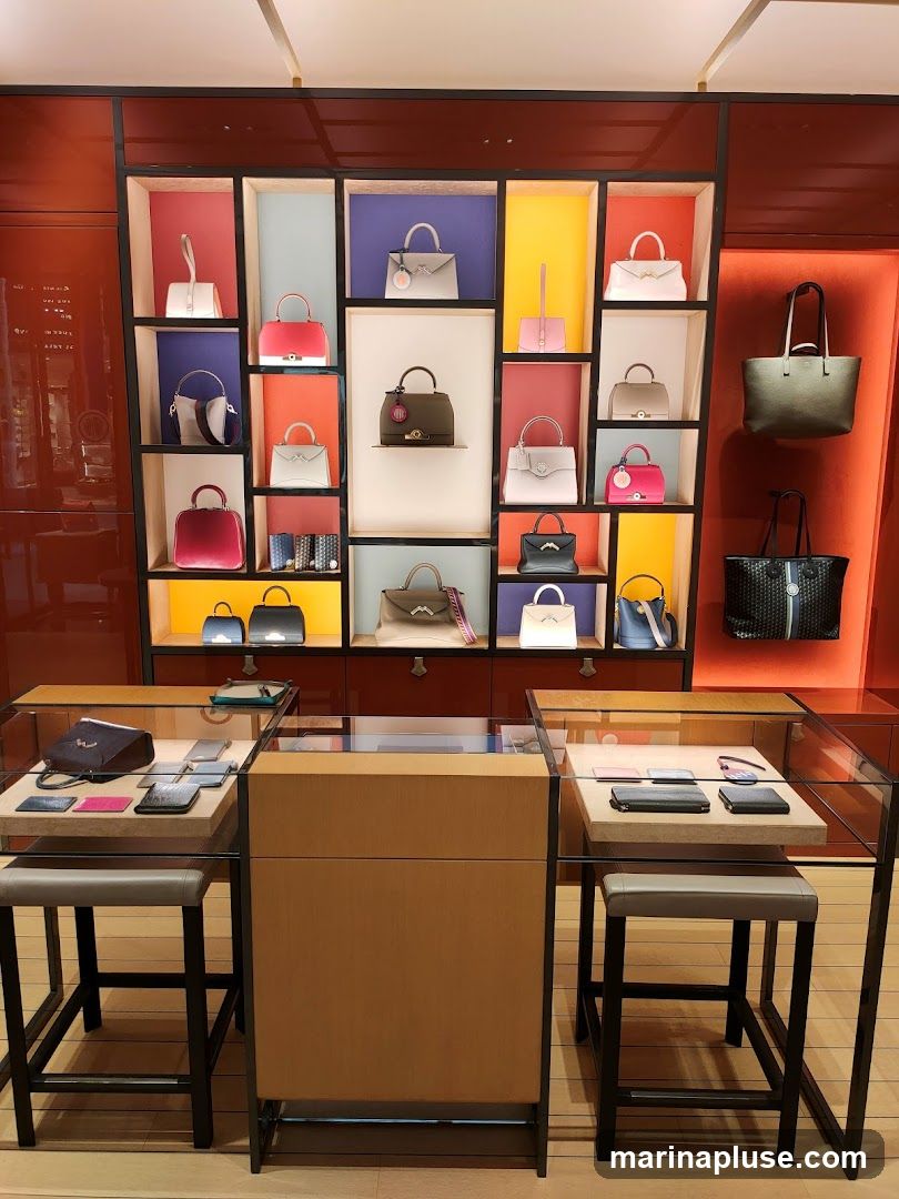 A retail display featuring a variety of handbags and small leather goods arranged on shelves and in glass cases. The handbags are in various colors and styles, including top-handle bags, crossbody bags, and clutches. Some bags have decorative charms or tags. The display is set against a backdrop of colorful shelves and a red accent wall.