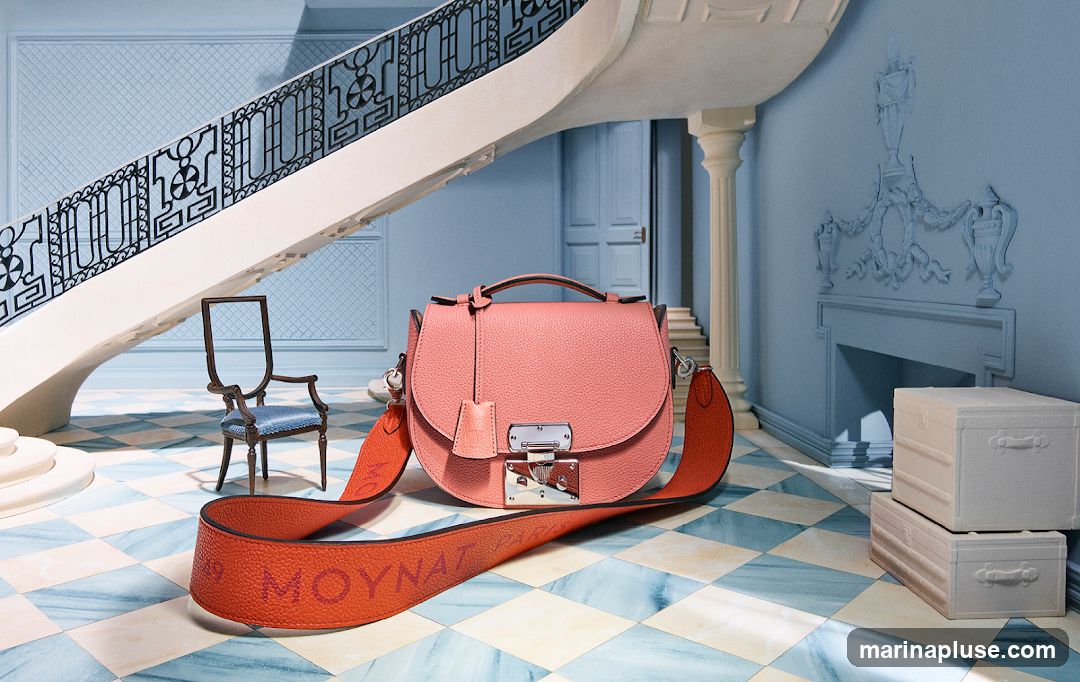 A pink textured leather handbag with an orange strap is displayed in a room with a grand staircase and blue walls. The strap has the embossed text "MOYNAT PARIS". A small antique chair and two stacked white trunks are also visible. The floor has a blue and white checkered pattern.