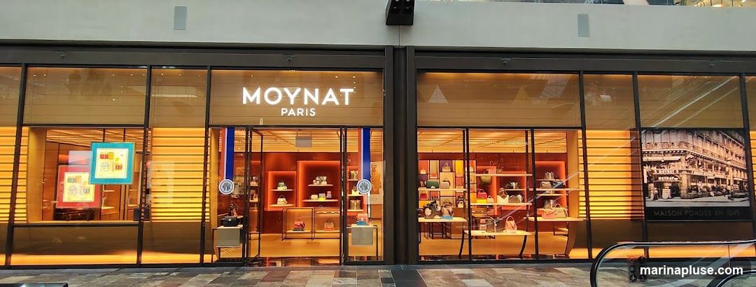The exterior of a Moynat Paris store. The storefront features large glass windows displaying handbags and accessories. The store's name, "MOYNAT PARIS," is prominently displayed in illuminated lettering above the entrance. A black and white historical image of a building with the text "MAISON FONDEE EN 1849" is visible on the right side of the storefront.