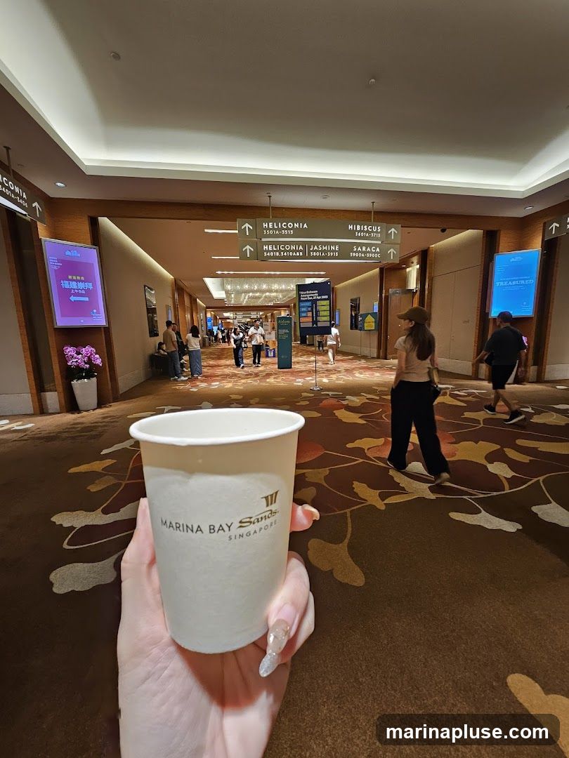 A hand holds a paper cup with the Marina Bay Sands logo. The cup is in the foreground, with a wide hallway stretching into the distance. Signage indicating directions to various areas like Heliconia, Hibiscus, and Saraca are visible on the ceiling. People are walking in the hallway, and digital screens display information. The floor is carpeted with a floral pattern.