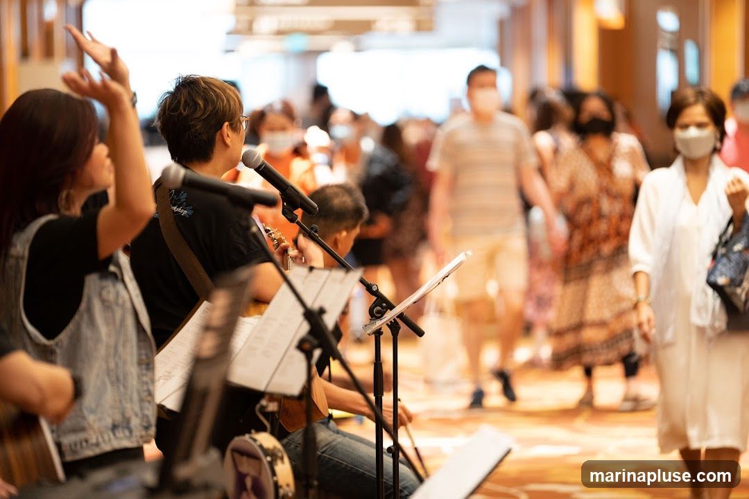 A group of musicians are performing in what appears to be a public indoor space, possibly a mall or convention center. In the foreground, a woman with her hands raised in worship or praise is visible. To her right, a musician plays a ukulele and sings into a microphone. Another musician is partially visible behind them. Music stands with sheet music are set up in front of the performers. In the background, a crowd of people, many wearing face masks, are walking by. The overall atmosphere suggests a lively event or gathering.