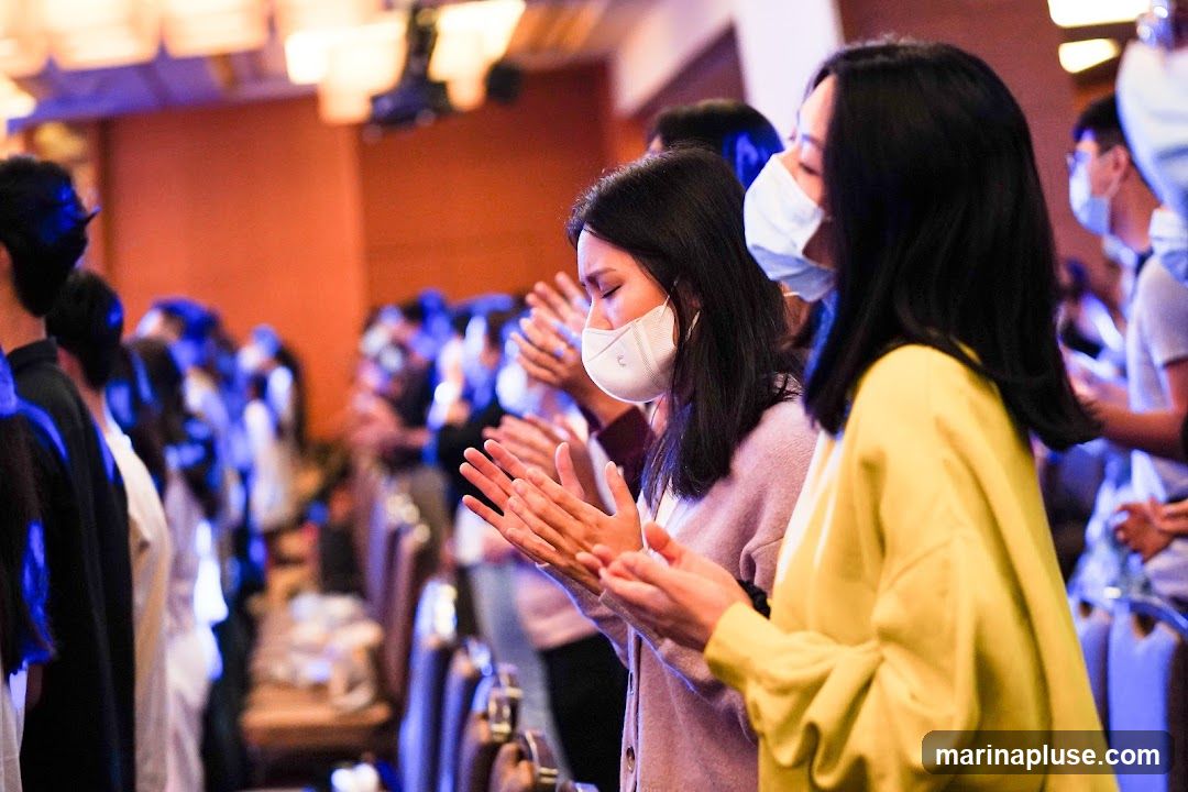 A group of people, mostly women, are shown from the chest up, clapping their hands. They are wearing face masks. The background is blurred, showing more people and a warm-toned wall with lights. The lighting is dim and atmospheric, with blue and purple hues present.