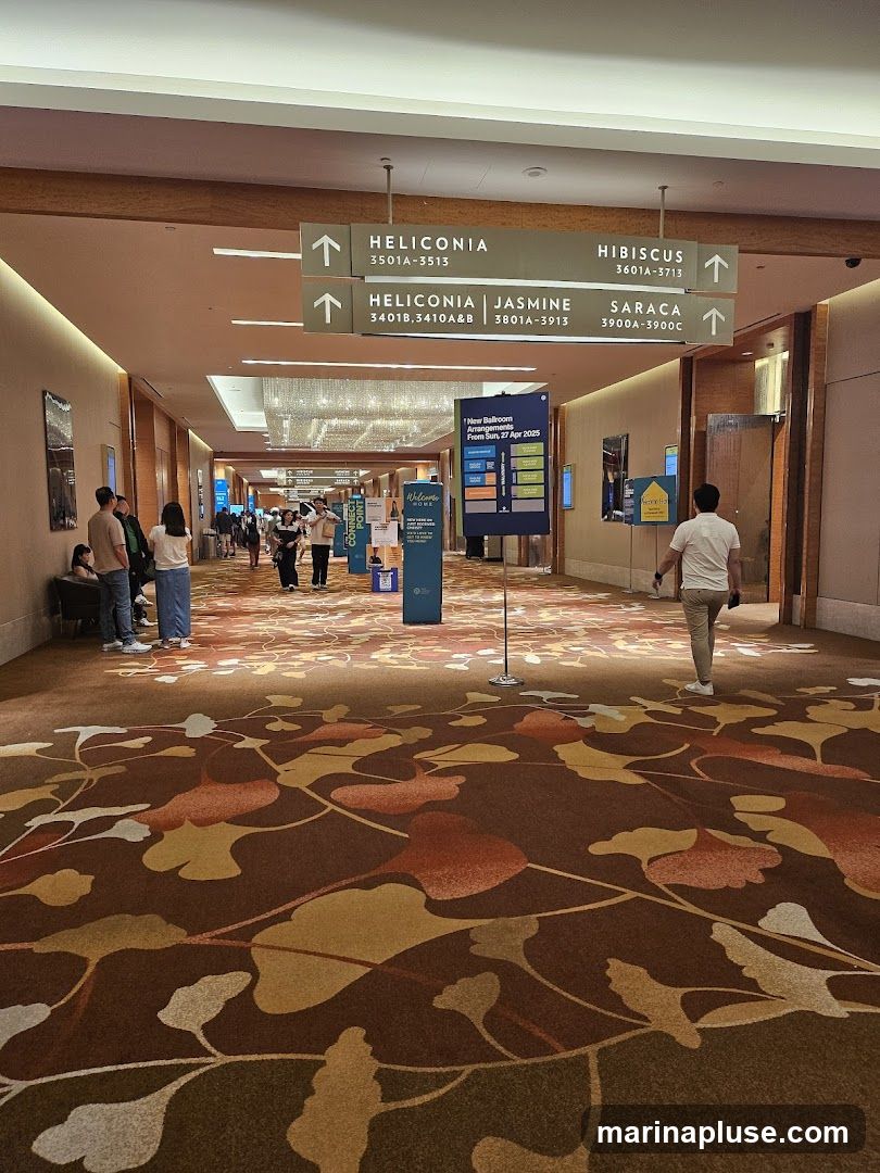 A wide hallway with directional signs pointing to different rooms like Heliconia, Hibiscus, Jasmine, and Saraca. The floor is covered with a patterned carpet. People are walking through the hallway, and there are informational displays.