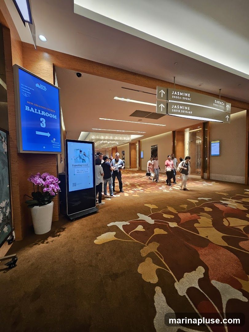 A wide hallway with a patterned carpet and directional signs. A digital display shows information about "New Creation Church Ballroom 3". Several people are walking through the hallway.