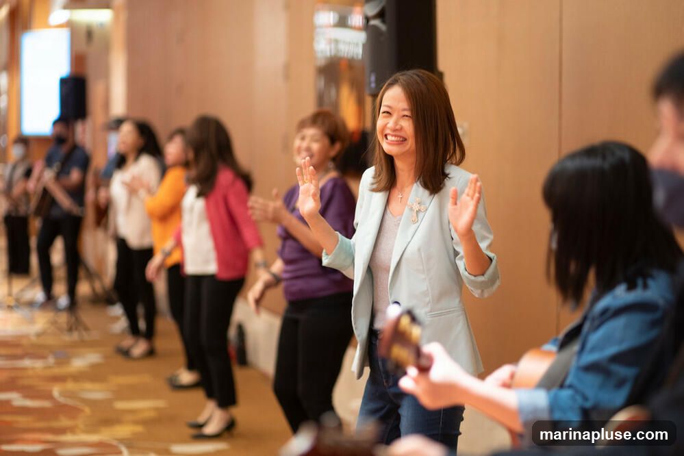 A group of people, mostly women, are standing in a line, clapping and singing. A woman in the foreground is wearing a light blue blazer and jeans, and she is smiling broadly with her hands raised. Another person in the foreground is playing a guitar. The background is blurred, but it appears to be an indoor event space with wooden walls.