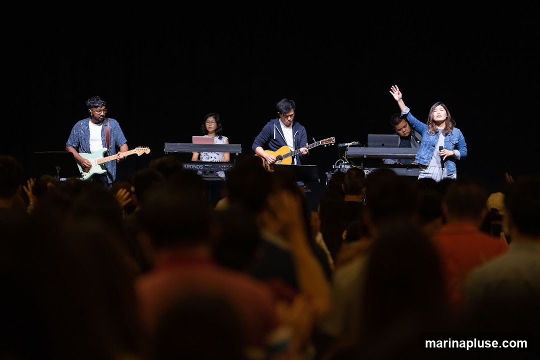 A band performs on a stage in front of a dark background. The band consists of a guitarist on the left, a keyboardist, another guitarist playing an acoustic guitar, and a singer on the right. A crowd is visible in the foreground, out of focus.
