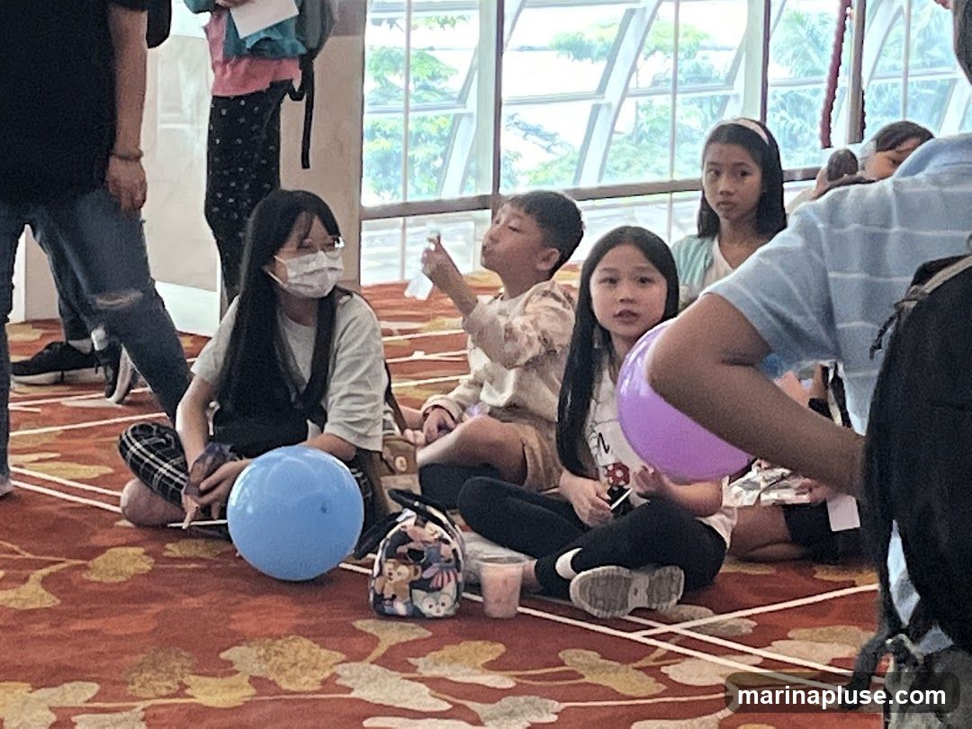 Several children are sitting on a red carpet with a gold pattern. Some are wearing masks. There are balloons and bags on the floor. In the background, there is a large window with a view of trees.