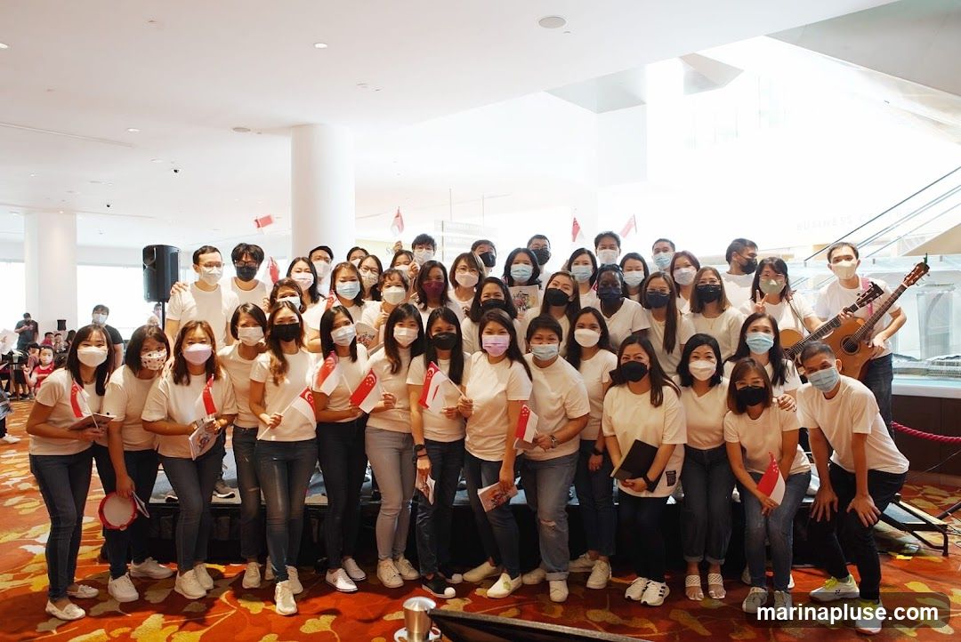 A large group of people, mostly women, are posing for a photo indoors. Many are wearing white t-shirts and jeans. Several individuals are holding small Singapore flags. Two people in the back right are holding guitars, and one is playing. A speaker is visible on the left. The background appears to be a modern indoor space with escalators and signage.