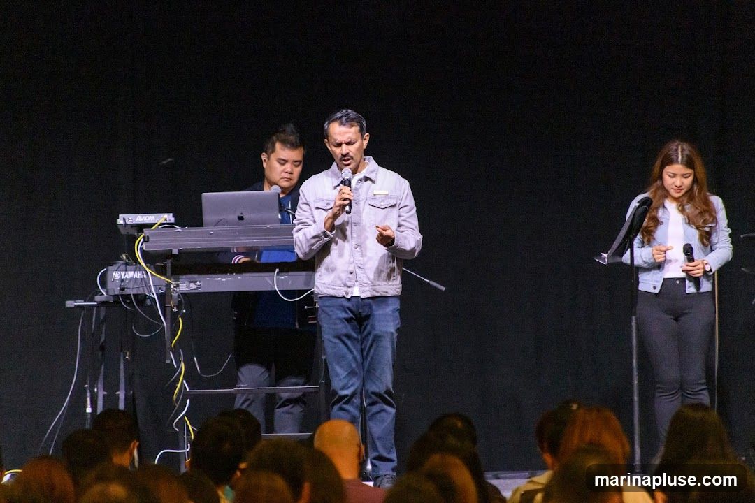 A man speaks into a microphone on a stage, wearing a light-colored denim jacket and jeans. To his left, a person operates a keyboard and laptop. To his right, a woman holds a microphone and stands near a music stand. An audience is visible in the foreground.