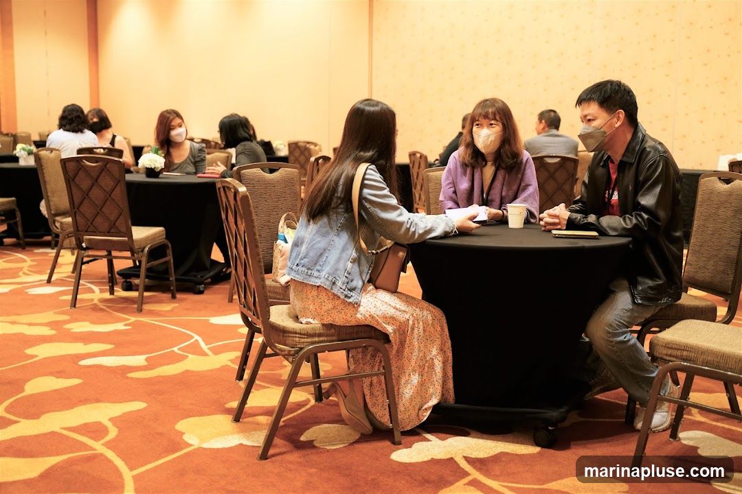 People are seated at round tables in a conference room, engaged in conversation. Most individuals are wearing face masks. The room has a patterned carpet and warm lighting.