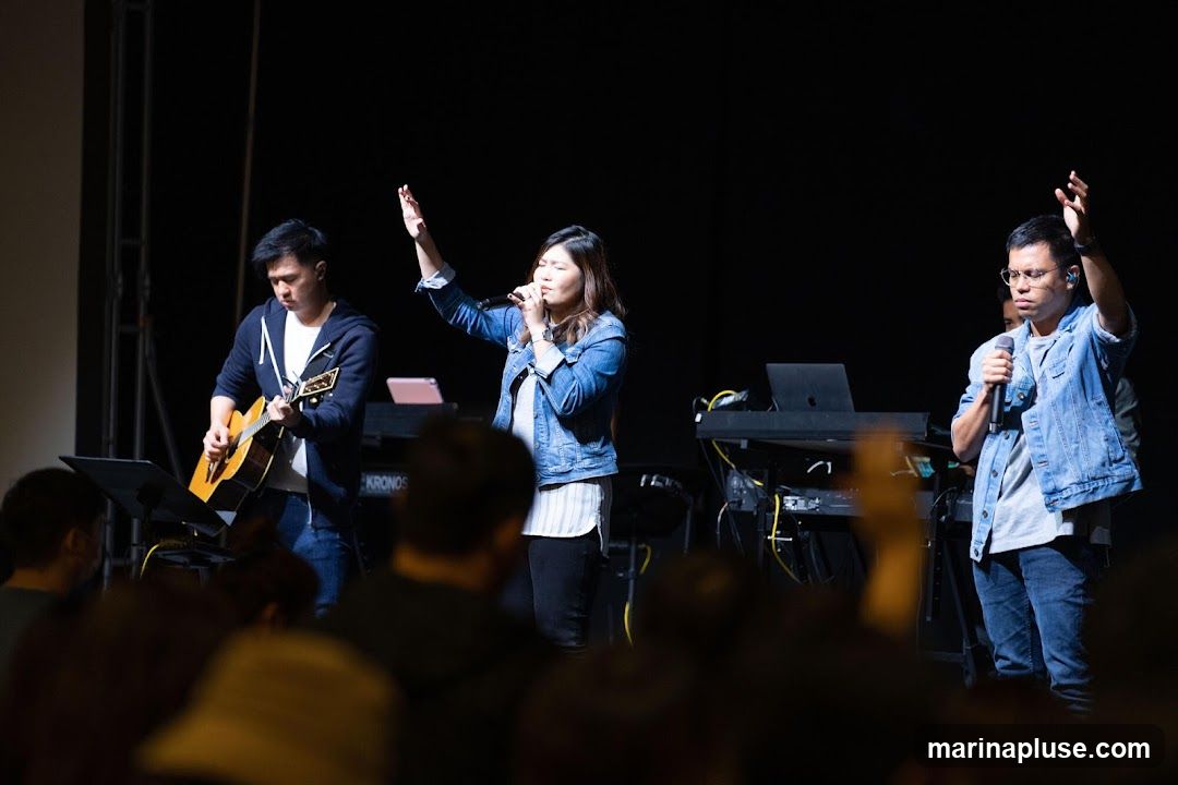 A worship band performs on a stage in front of a dark background. A man on the left plays an acoustic guitar. A woman in the center sings into a microphone, with her hand raised. A man on the right also holds a microphone and has his hand raised. Behind them are keyboards and other musical equipment. An audience is visible in the foreground, out of focus.