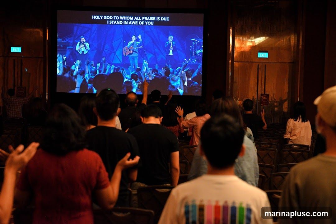 A group of people are seated in a room, facing a large screen. The screen displays lyrics to a song, "HOLY GOD TO WHOM ALL PRAISE IS DUE I STAND IN AWE OF YOU," and an image of a band performing. Some people in the audience have their hands raised. An "EXIT" sign is visible on a door in the background.