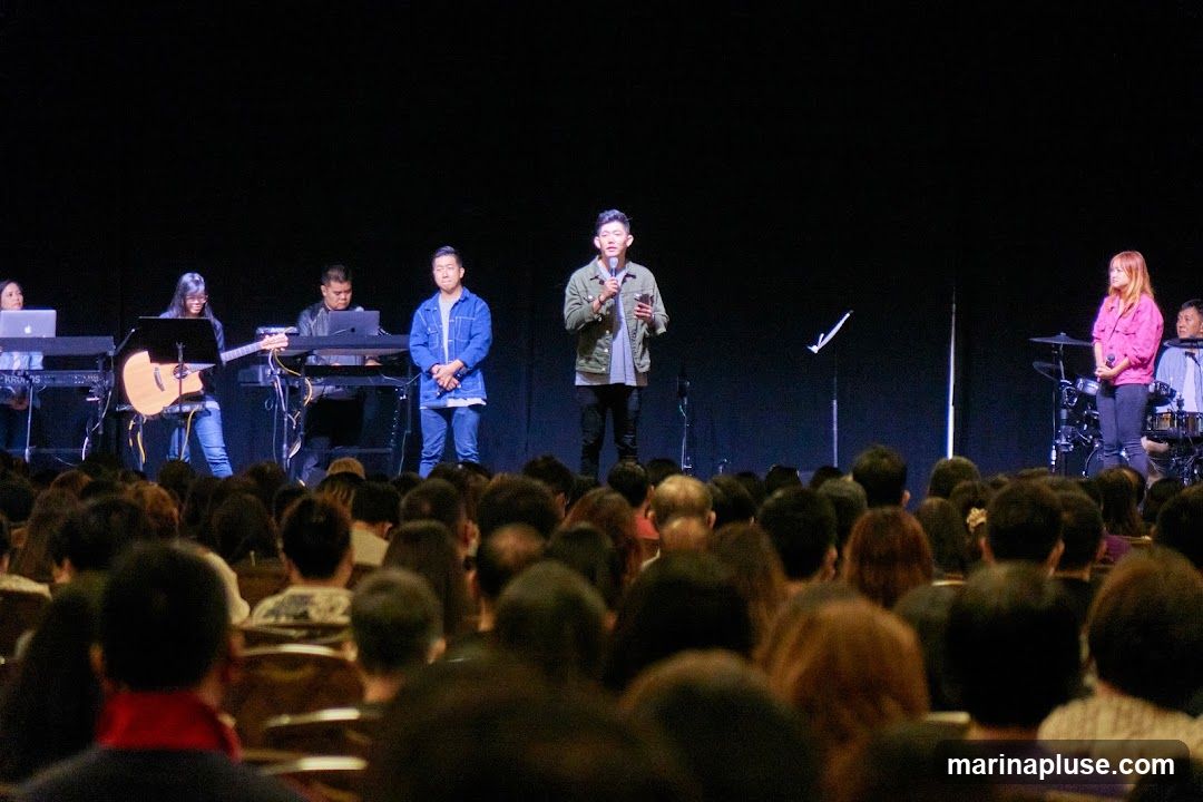 A live performance is taking place on a stage in front of a large audience. On stage, a band is set up with keyboards, a guitar, and drums. Several people are on stage, including musicians and two men who appear to be speakers or hosts. One man is holding a microphone and speaking, while the other stands beside him. A woman in a pink jacket is also visible on the right side of the stage, near the drums. The audience is seated in a dark room, facing the stage.