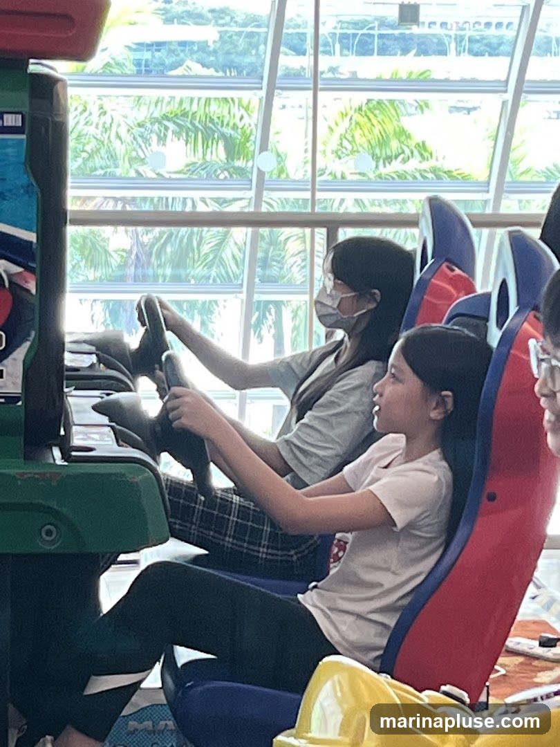 Two young girls are seated in arcade racing game machines, each holding a steering wheel. The girl in the foreground is wearing a light-colored t-shirt and dark leggings, while the girl behind her wears a grey t-shirt and plaid pants, and a face mask. They are positioned in front of a large window with a view of palm trees and buildings. The arcade machines have colorful seats and controls.