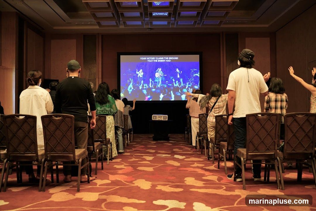 A group of people stand in a conference room, facing a large screen that displays lyrics and an image of a band performing. The room has a red carpet with a floral pattern and rows of chairs. The people appear to be engaged in a worship or concert-like event.