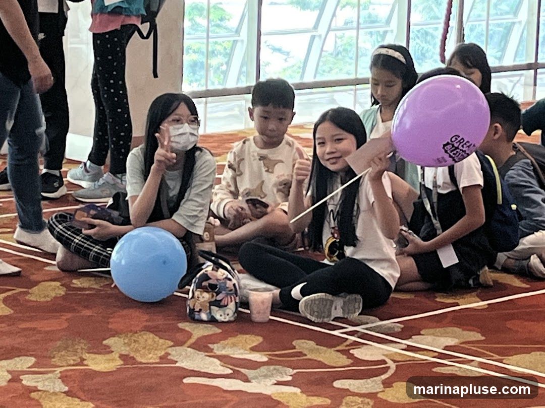 A group of children are sitting on a patterned carpet, some holding balloons. One girl in the foreground is holding a purple balloon with text that appears to say "GREAT EASTERN". Another girl to her left is wearing a mask and holding up two fingers in a peace sign. There are two balloons on the floor, one blue and one purple. The children are dressed casually.