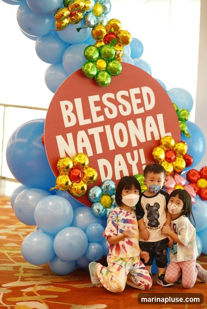 Three children, wearing masks, pose in front of a balloon arch and a red circular sign that reads "BLESSED NATIONAL DAY!". The balloon arch is made of light blue balloons and decorated with foil balloons shaped like flowers in gold, green, and blue. The children are dressed casually, with two girls and one boy. The floor has an orange patterned carpet.