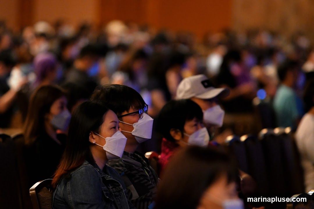A crowd of people, mostly wearing white face masks, are seated in a dimly lit room. The focus is on a few individuals in the foreground, with the rest of the audience blurred in the background. The overall impression is of a gathering or event where mask-wearing is prevalent.
