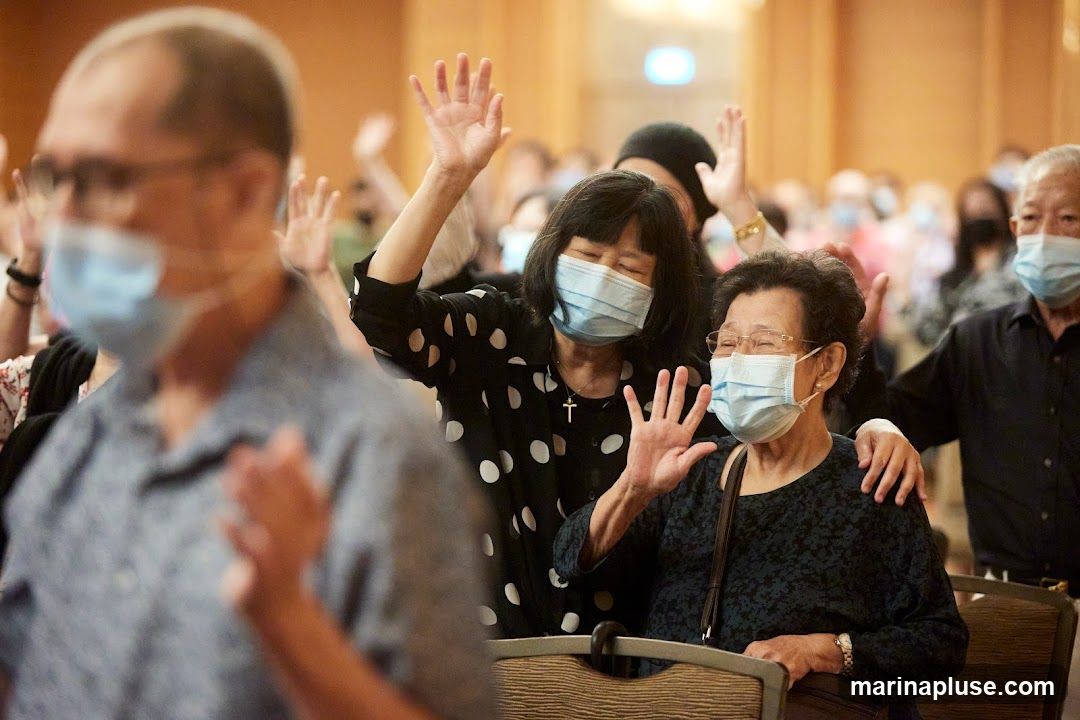 A group of people, mostly elderly, are seen with their hands raised in the air. Most of them are wearing face masks. The setting appears to be an indoor gathering, possibly a religious service or a conference. The focus is on the individuals in the foreground, with others blurred in the background.