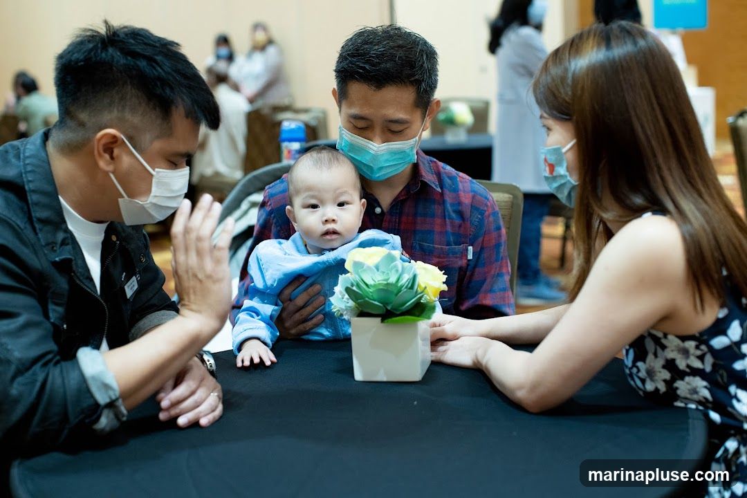 A family, including a baby, is seated at a table. The adults are wearing face masks. A small floral arrangement is on the table. In the background, other people are visible, some also wearing masks.