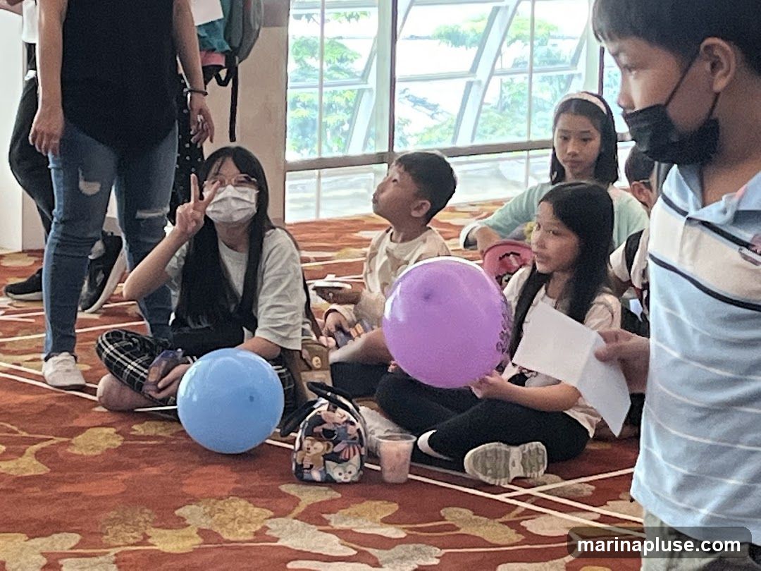 A group of children are sitting on a patterned carpet, some with balloons. One girl in the foreground is wearing a mask and holding up two fingers in a peace sign. Another child to her right is holding a piece of paper. In the background, large windows allow natural light to enter the room.