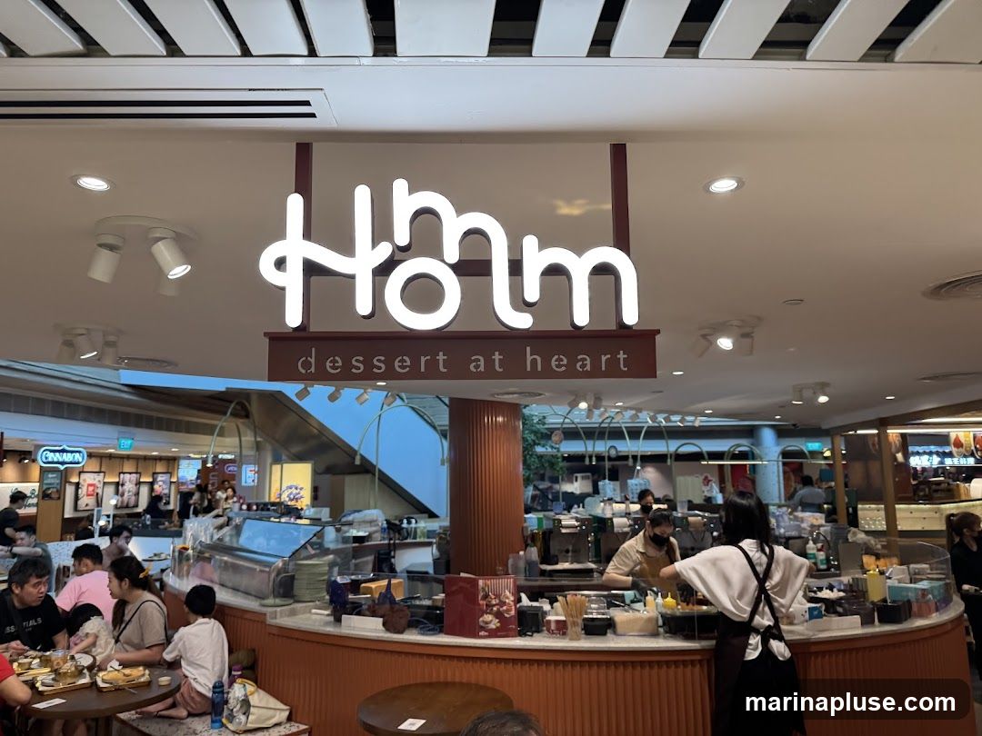 An interior view of a dessert shop with a prominent neon sign that reads "Hmm dessert at heart." Several customers are seated at tables, and staff members are visible behind the counter preparing orders. The shop appears to be located within a mall, with escalators and other storefronts visible in the background.