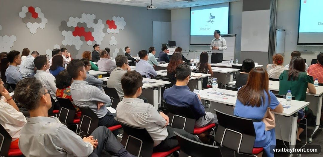 A group of people are seated at tables in a conference room, facing a projector screen. A man is standing and presenting. The walls are decorated with a hexagonal pattern.