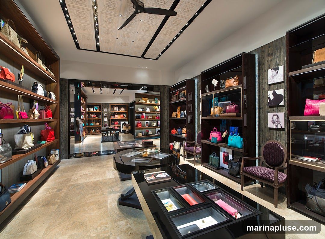 A well-lit, upscale boutique interior showcasing a wide selection of designer handbags on shelves and display cases. The store features polished floors, dark wood shelving, and modern lighting. Several seating areas with upholstered chairs and benches are visible, along with decorative artwork on the walls.