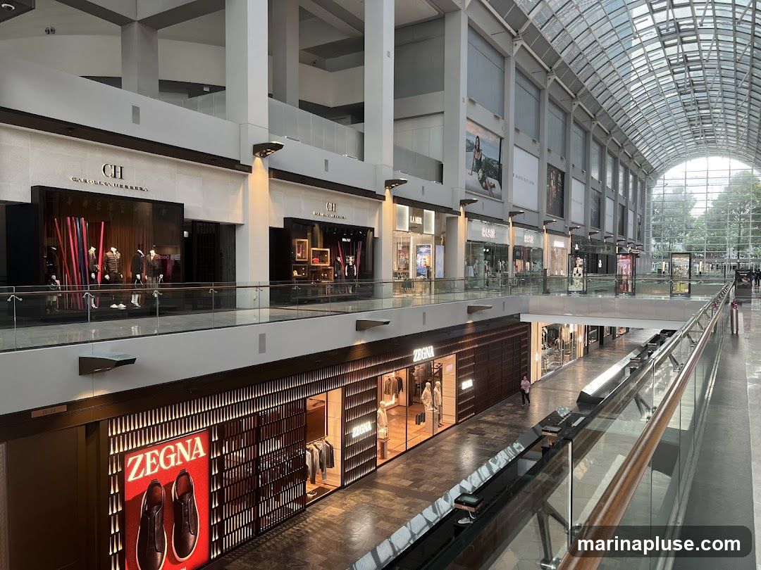An interior view of a modern shopping mall with multiple levels and a glass ceiling. Several high-end retail stores are visible, including Carolina Herrera and Zegna, with their storefronts and displays. The mall features a contemporary design with clean lines and ample natural light filtering through the arched glass roof.