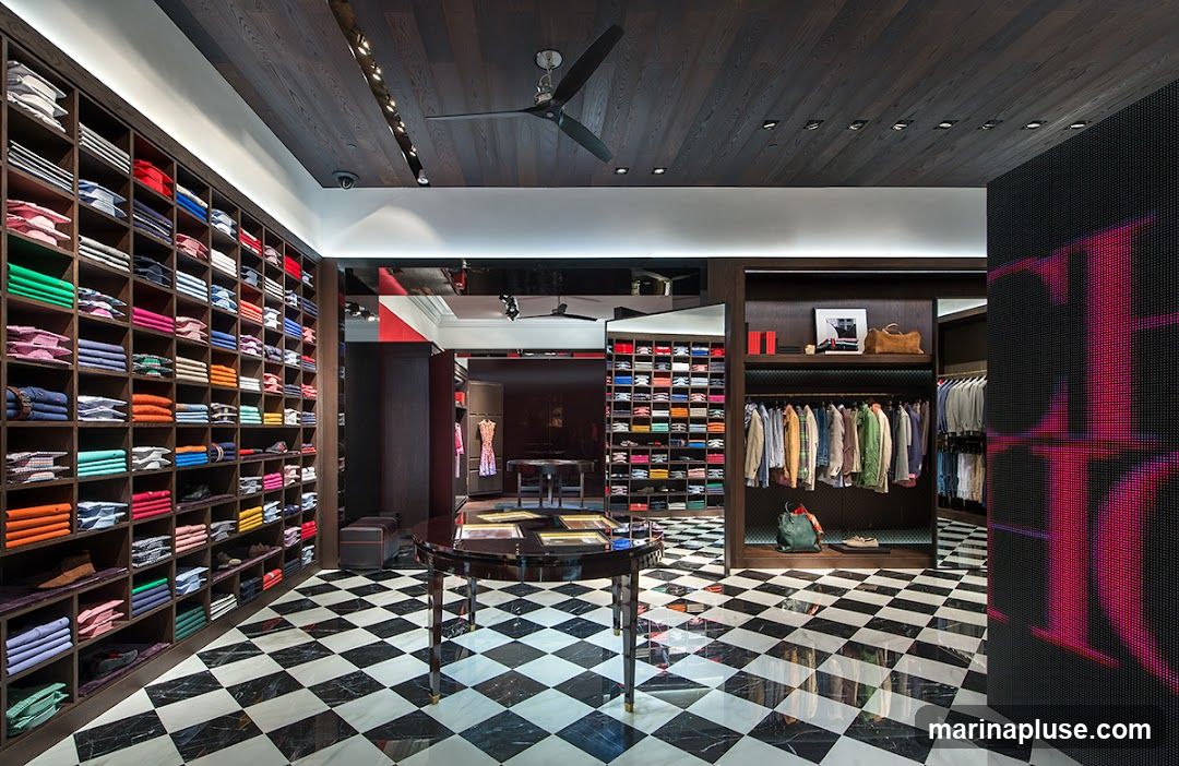 A well-lit, modern clothing store interior with a black and white checkered marble floor. Floor-to-ceiling shelves on the left are filled with neatly folded colorful shirts. In the center, a display table with a glossy black surface. To the right, racks of clothing are displayed, and a large digital screen on the far right shows a pink logo. The ceiling is dark wood with recessed lighting and a large black ceiling fan.
