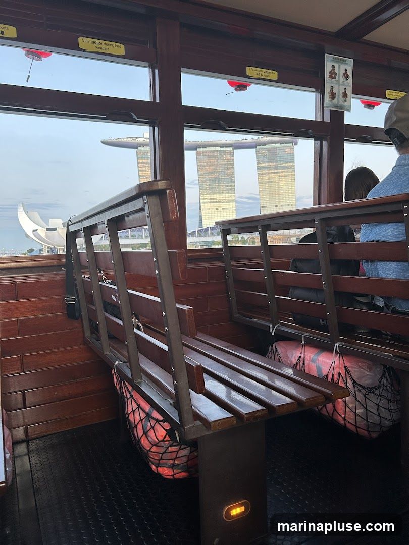 The image shows the interior of a boat with wooden benches and railings. Through the windows, the iconic Marina Bay Sands hotel in Singapore is visible in the distance, along with the ArtScience Museum. There are safety instructions and life jackets visible inside the boat.